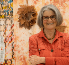 The textile artist Lesley Turner poses for a headshot in front of her inspiration wall, wearing a burnt orange collared shirt, glasses and a warm smile